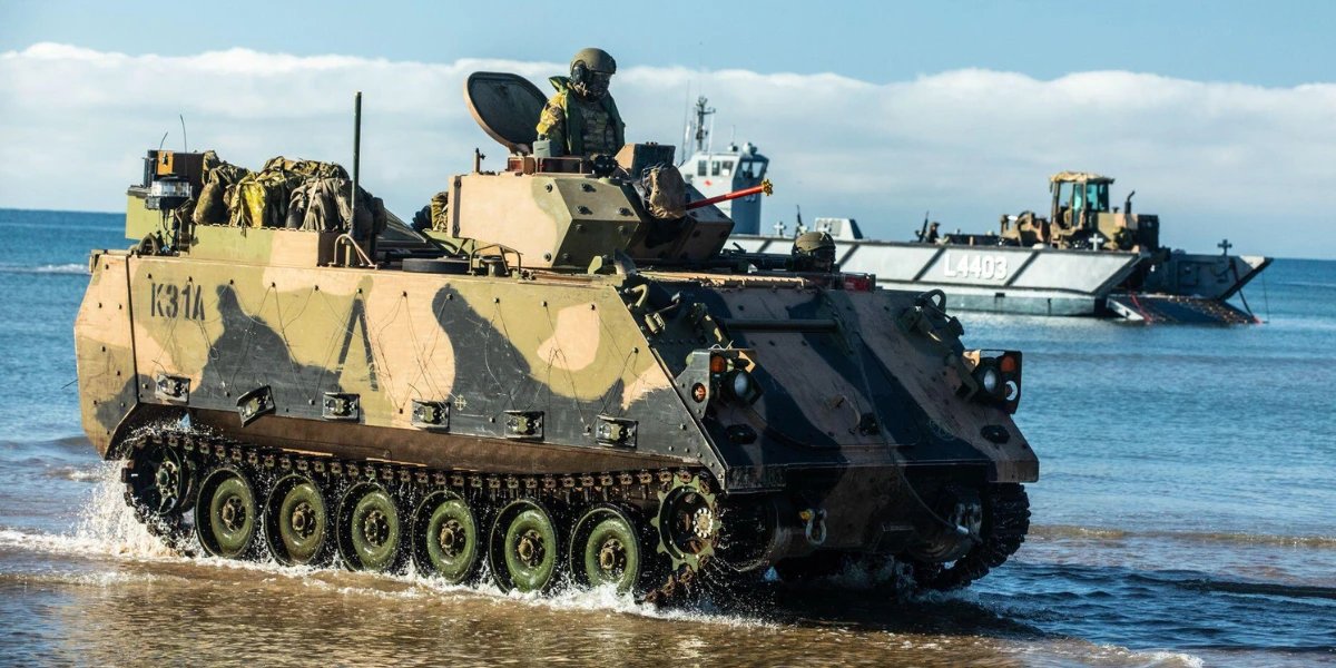 An Australian Army M113 armoured personnel carrier drives through shallow water on a beach, with a landing craft behind it