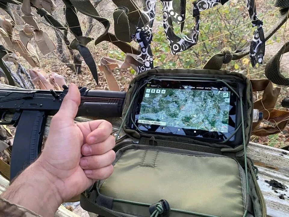 A soldier gives a thumbs-up next to a rugged tablet in a pouch showing the Kropyva map application, under camouflage netting