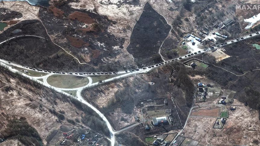 Satellite view of a long column of military vehicles on a road through burnt fields and a village