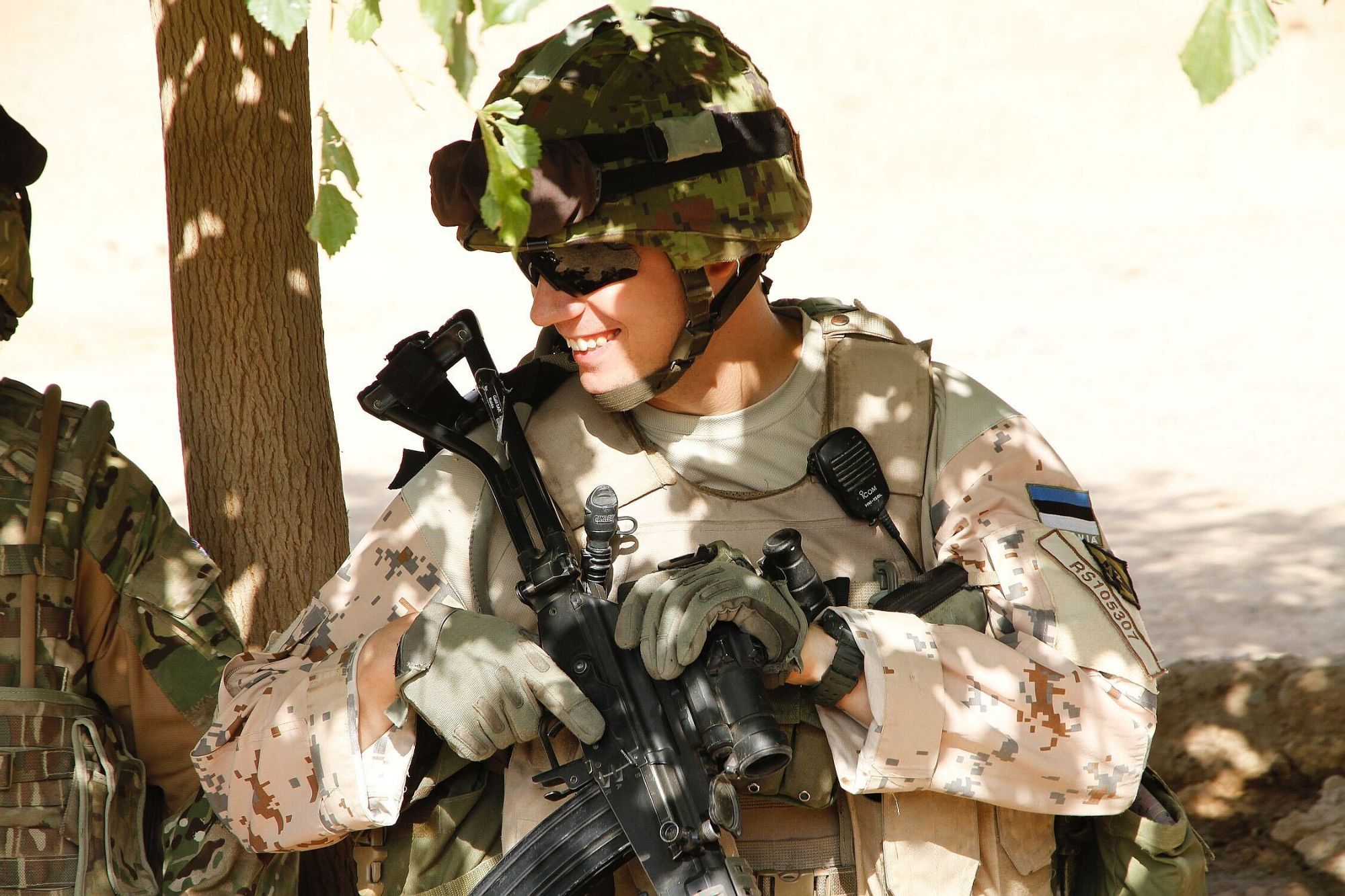 Raido Saremat, smiling, in Estonian desert uniform and helmet with his rifle, standing in the shade of a tree